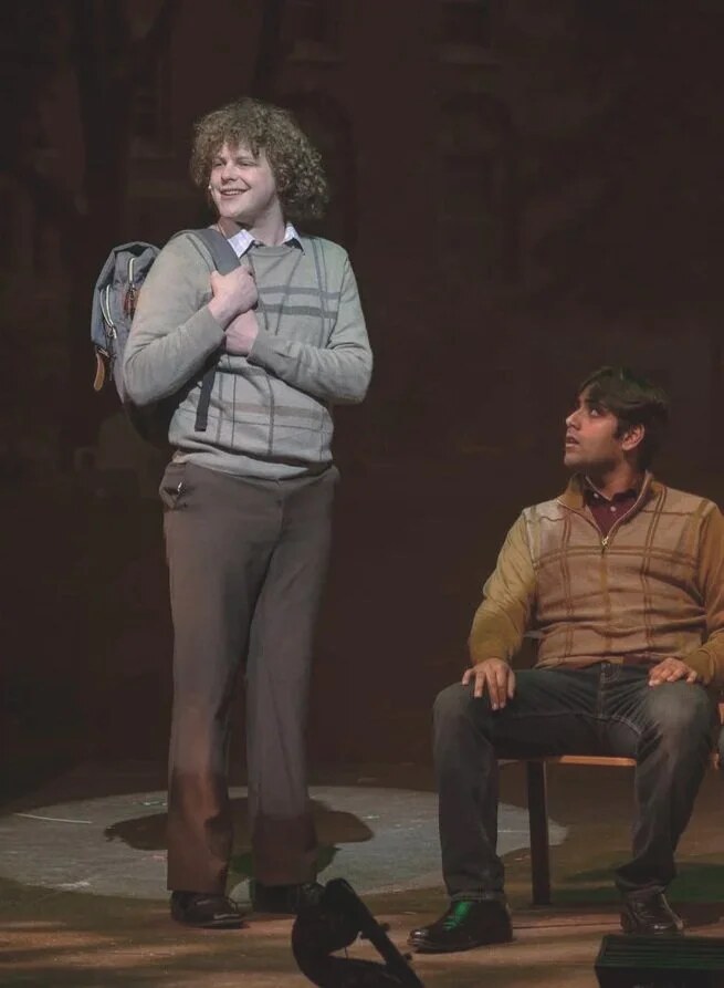 A curly-haired performer in a gray sweater smiles and holds a backpack strap, while a seated performer looks up from a chair.