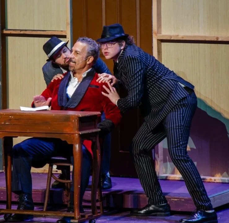 Two performers in fedoras, one in a pinstripe suit, lean in over a seated performer in a red velvet jacket at a small wooden desk.