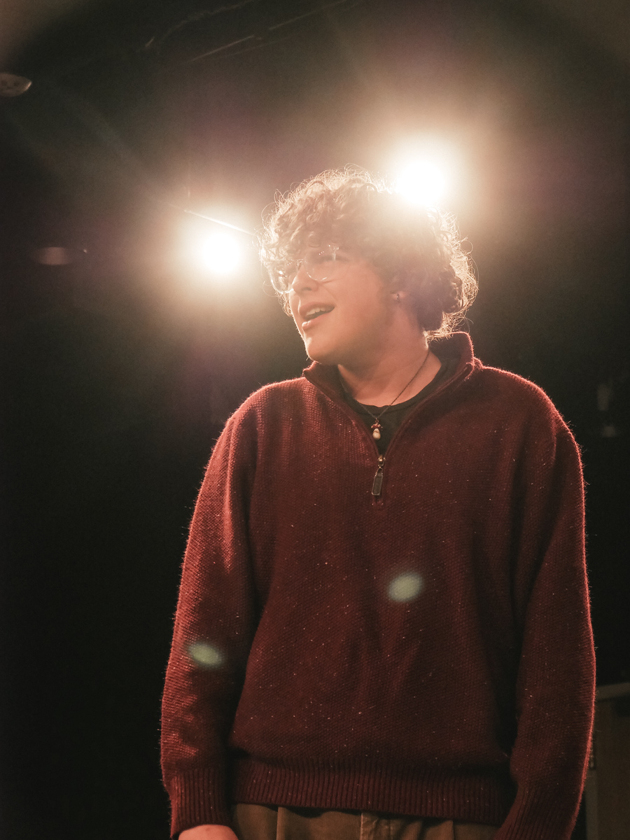 A curly-haired performer in a burgundy sweater sings under bright backlights.